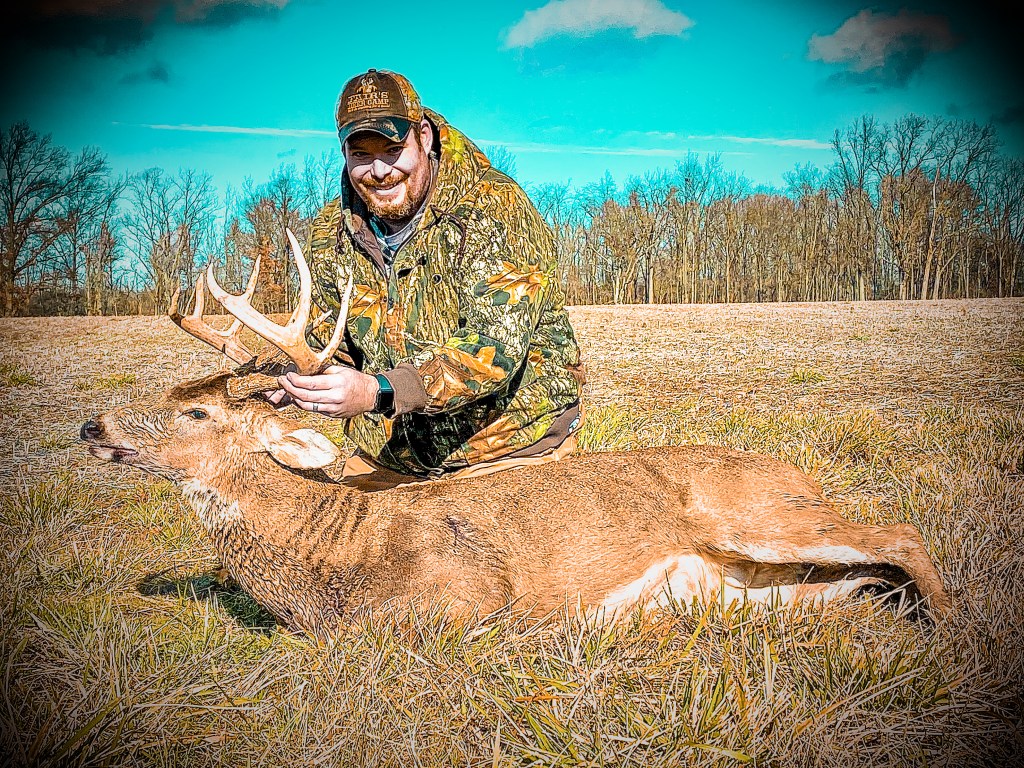 A man in camouflage attire poses beside a freshly hunted deer in an open field with trees in the background.